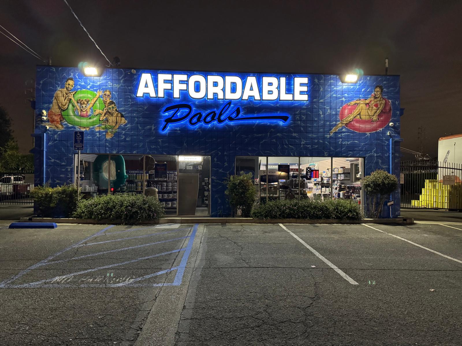 Affordable Pools storefront at night with illuminated white channel letters and a blue backlit script over a pool mural